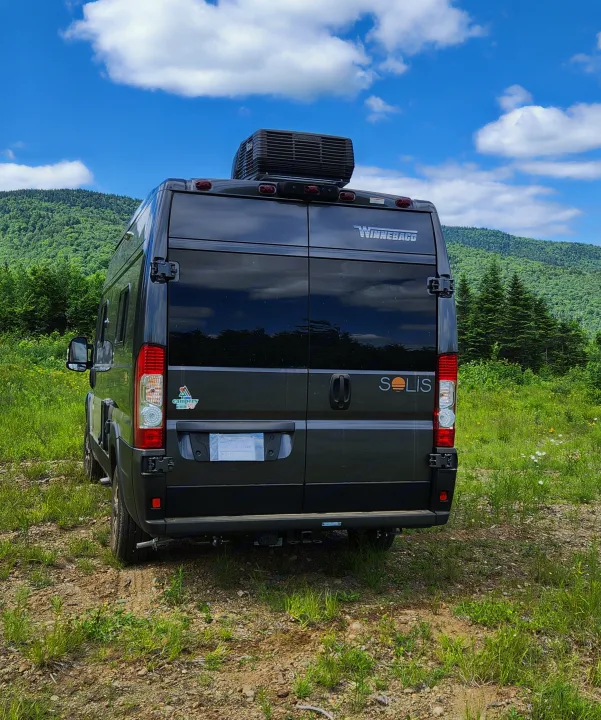 Rear view of a dark Winnebago Solis **rv rental** parked on a grassy path with green mountains and a blue sky.. Winnebago Solis 2024