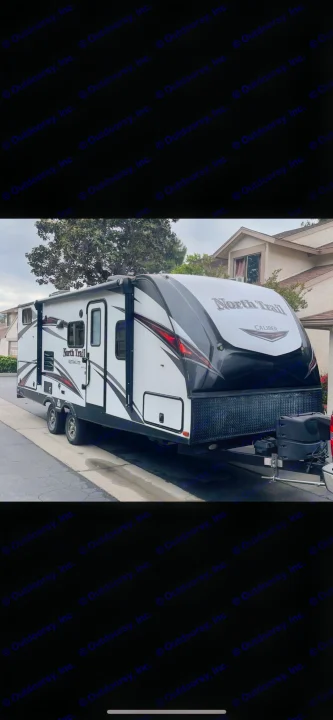 North Trail travel trailer RV for rent. This white and black RV with tandem axles is parked on a paved residential street.. Heartland RVs North Trail 2019