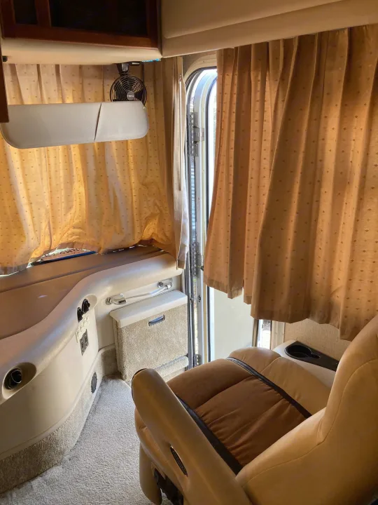 Inside an RV rental: a warm interior with beige and brown seating, textured curtains, a small fan, and light carpet.. Fleetwood Expedition 2003