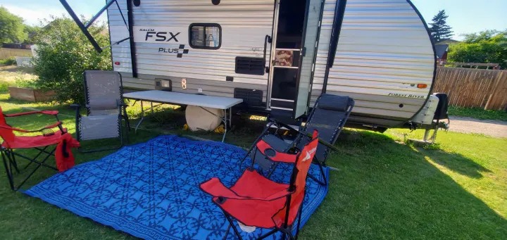 A bright side view of an RV rental with an outdoor camping setup on green grass. Features a blue mat, red and grey chairs, and a white table.. Forest River Salem FSX 2019