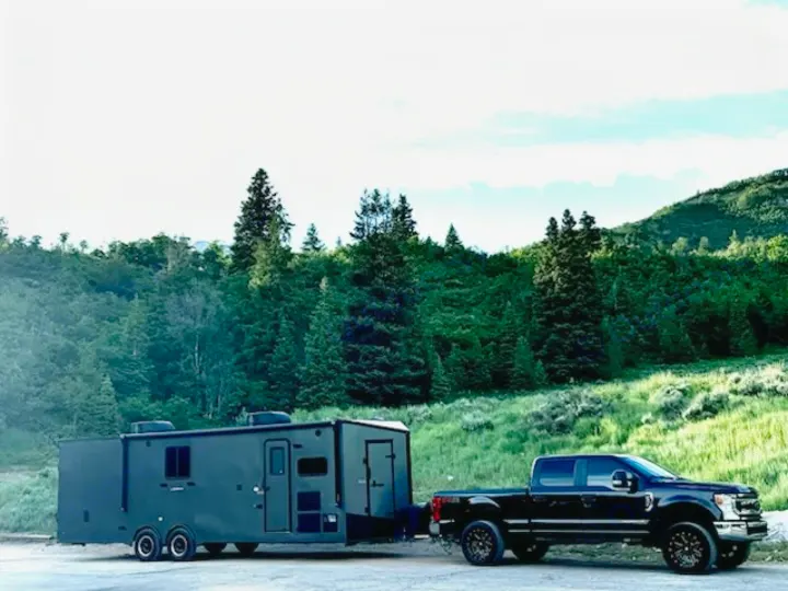Black truck towing a dark, robust travel trailer in a lush mountain landscape. Ideal for a spacious rv rental experience.. Stealth Nomad 28FK 2022