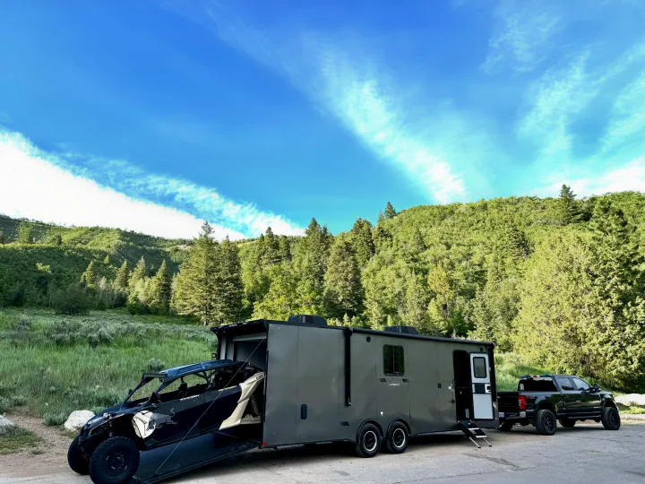 Ultimate adventure RV rental setup: a large toy hauler trailer with an off-road UTV and heavy-duty pickup truck by mountains.. Stealth Nomad 28FK 2022