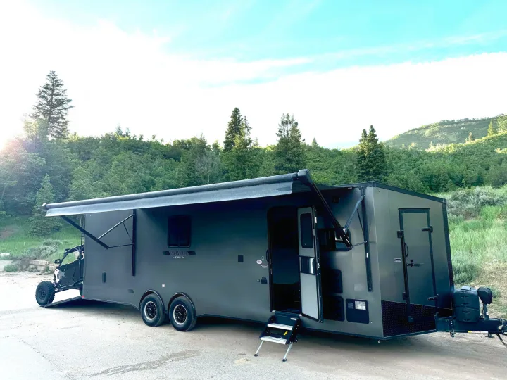 Adventure-ready dark gray toy hauler RV rental with UTV, awning out, nestled in mountains.. Stealth Nomad 28FK 2022