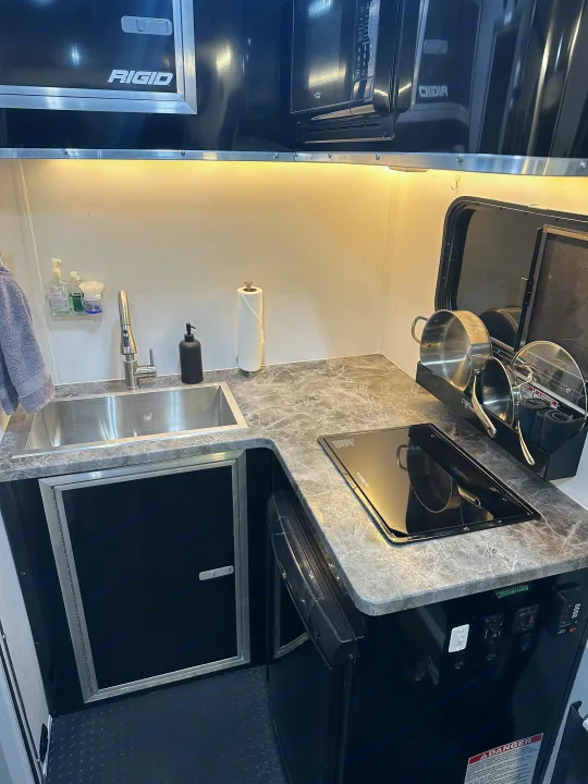 Compact RV rental kitchenette features a stainless sink, Dometic induction stove, microwave, and sleek black cabinets.. Stealth Nomad 28FK 2022
