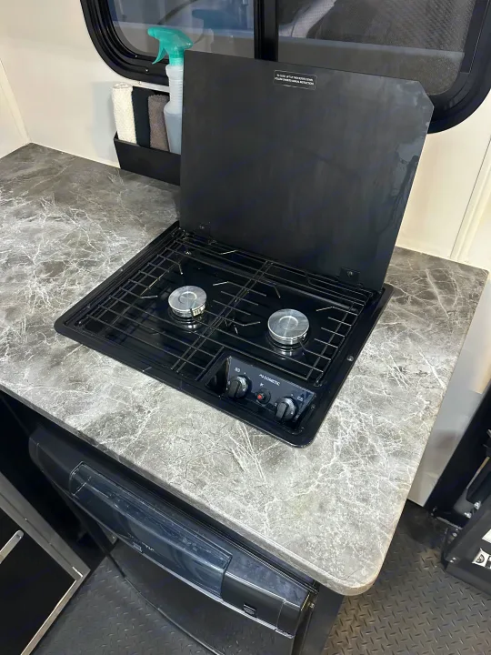 An RV rental kitchen features a Dometic two-burner stove on a marble-look countertop, with a lift-up cover for travel.. Stealth Nomad 28FK 2022