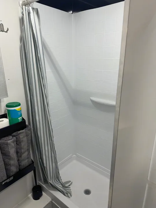 Clean, modern RV rental shower with striped curtain, shelf, and organized amenities like towels & wipes.. Stealth Nomad 28FK 2022