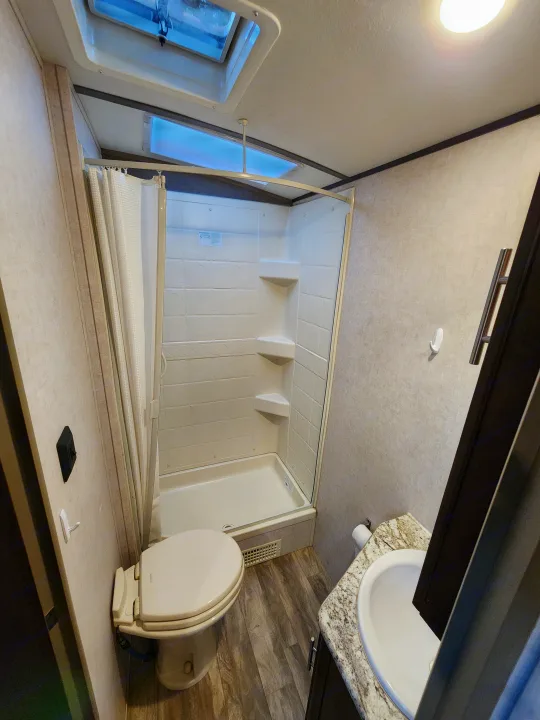 Compact RV rental bathroom featuring a toilet, spacious shower with curtain, sink vanity, and overhead skylight.. Forest River Surveyor Legend 2019