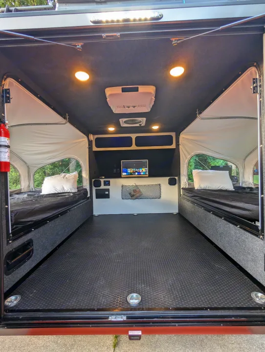 Interior of an RV rental, showing two beds with white pillows, a central floor, and a rear wall with a TV & speakers.. Intech Flyer Explore 2022