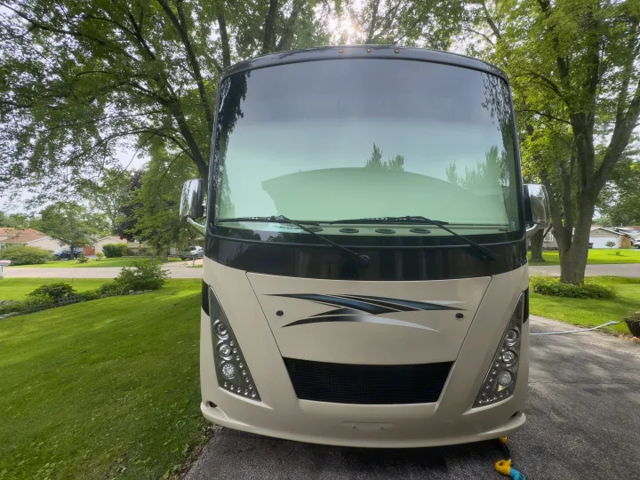 Front of a tan motorhome, possibly an RV rental, parked in a driveway with trees and houses in the background.. Thor Windsport 2020
