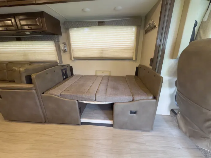 Inside an RV rental: a beige upholstered seating area converted into a bed, with overhead cabinets and window blinds.. Thor Windsport 2020