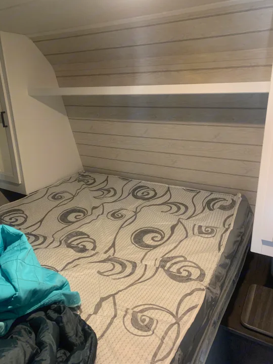 View of a bed with a grey patterned mattress inside an RV rental, featuring a practical overhead shelf and side cabinetry.. Keystone RV Hideout 2023
