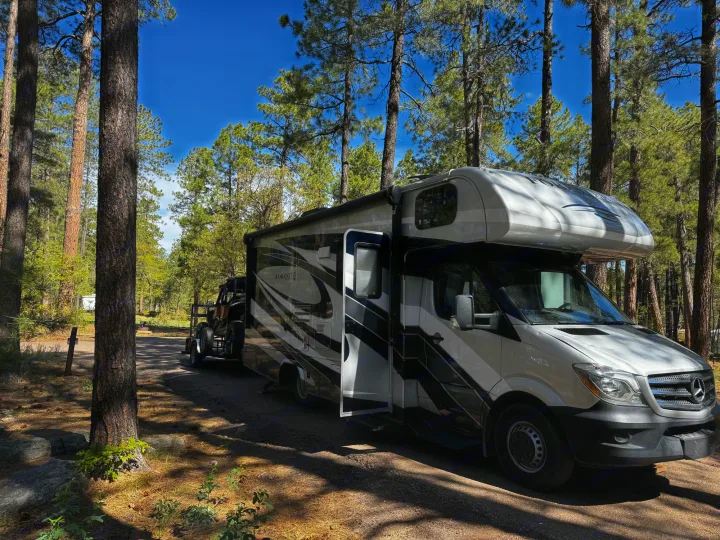White and black RV rental parked in a sunny forest campsite, towing a UTV.. Forest River Mercedes 2019