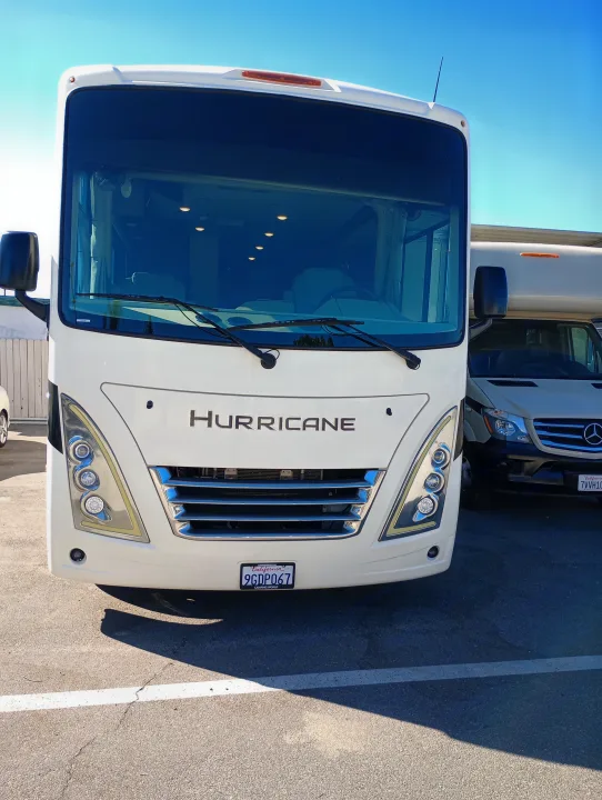 A white Hurricane RV rental is prominently parked on an asphalt lot in California, with a smaller van visible to its right.. Thor Motor Coach Hurricane 2022