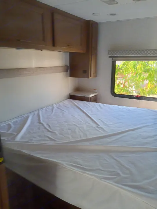 Cozy RV rental bedroom features a bed with white sheet, wood cabinets, nightstand, and window with foliage.. Thor Motor Coach Hurricane 2022