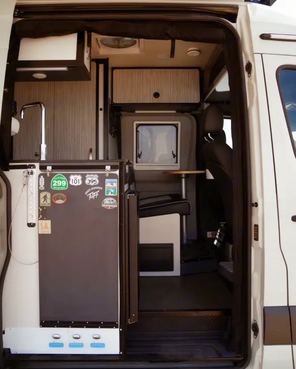 Step into this RV rental camper van, featuring a modern kitchenette, dining area, and travel stickers on the fridge door.. Winnebago Revel 2022