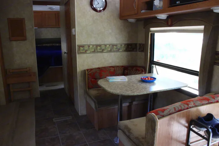 Cozy RV rental interior: a dinette with table, patterned bench seats, books, and cherries. Shoes stored below. Bedroom visible beyond.. Jayco Jay Flight G2 2008