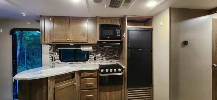 Interior of an RV rental kitchen, featuring a sink, microwave, stove, and refrigerator, with a view of trees outside.. Aspen Trail Aspen Trail Trailer 2019