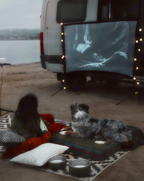RV rental getaway: Person and dog enjoy an outdoor movie by the water next to their camper van.. Winnebago Revel 2022