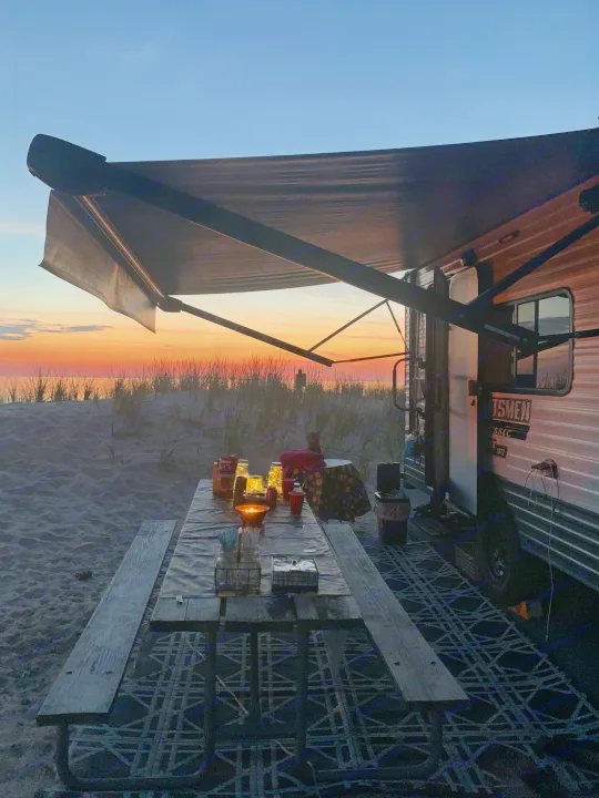 Sunset beach camping with a cozy RV rental setup. Enjoy outdoor dining by the water's edge at dusk.. KZ Sportsmen classic 160QB 2021