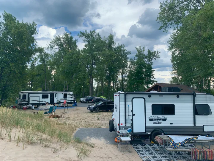 Two RV rental campers at a sandy site by the woods. Outdoor setup with tables, chairs, and a fire pit under clouds.. KZ Sportsmen classic 160QB 2021