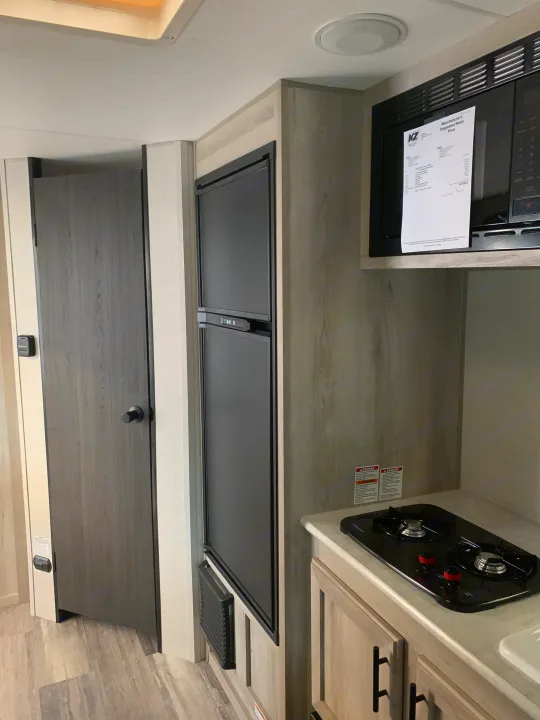 RV rental interior: compact kitchen with fridge, microwave, and two-burner cooktop, next to a closed door.. KZ Sportsmen classic 160QB 2021