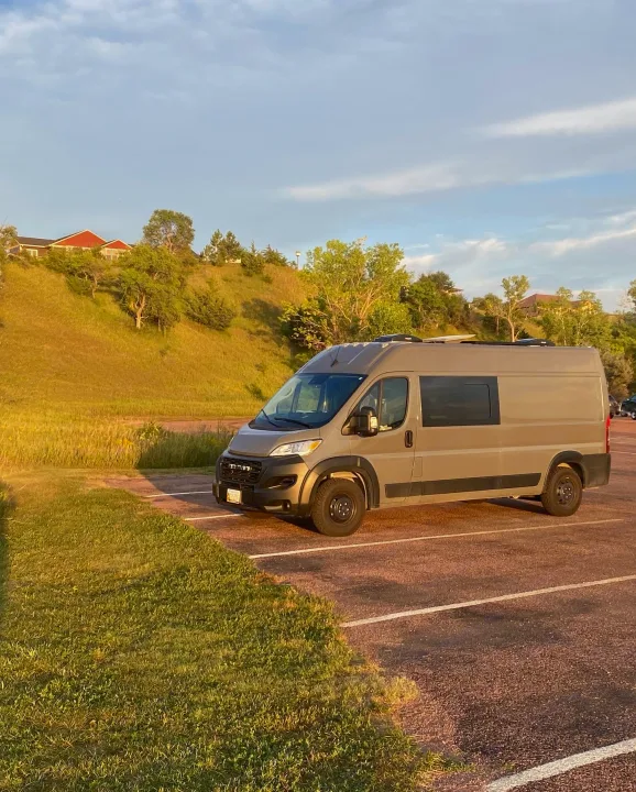 Modern grey camper van, perfect for an RV rental adventure, parked on asphalt beside sun-kissed hills and houses.. RAM Promaster 2300 2023