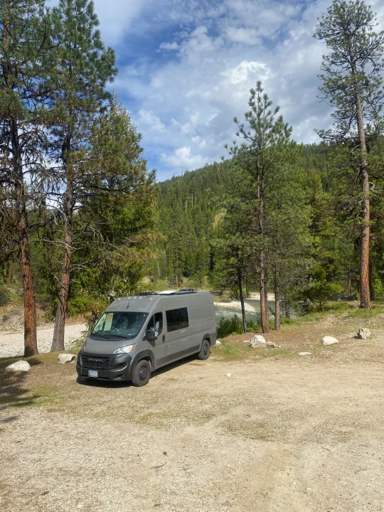 Gray camper van RV rental parked by a river in a mountainous forest setting under a cloudy blue sky.. RAM Promaster 2300 2023