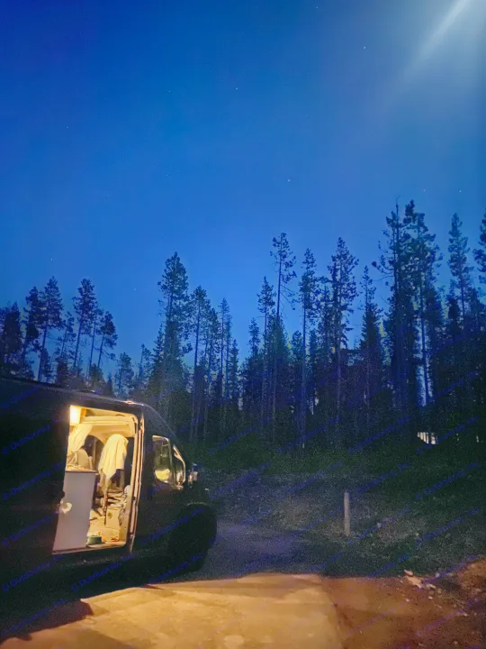 Night view: an RV rental with its interior lights glowing, nestled in a dark forest under a sky full of stars.. RAM Promaster 2300 2023