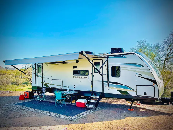 A white travel trailer (RV rental) under a blue sky, set up with an awning, chairs, and table for desert camping.. Keystone RV Passport GT Ultra Lite 2023