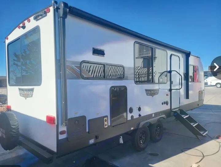 This white and grey Wildwood travel trailer, ideal for an RV rental, is parked outdoors, featuring its side door steps and rear.. Forest River Wildwood X-Lite 2024