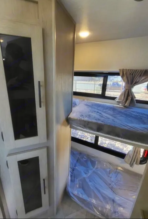 RV rental interior: light wood cabinets on left, two bunks on right, one still in plastic wrap. Bright and clean.. Forest River Wildwood X-Lite 2024