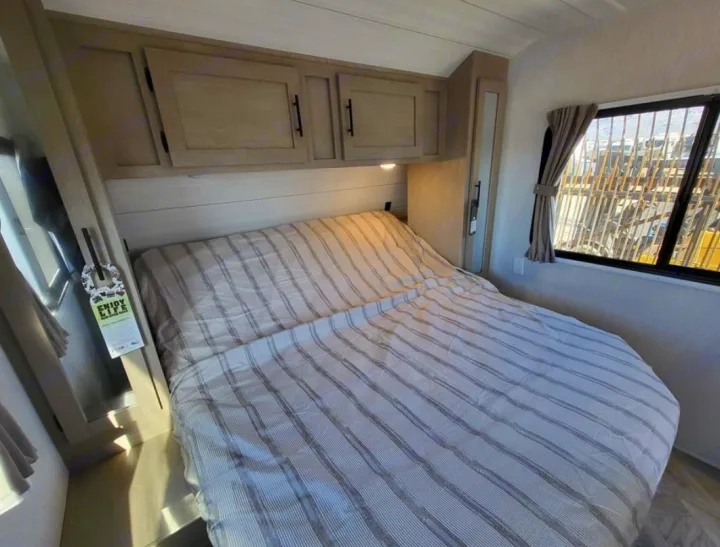 Compact RV rental bedroom featuring a comfortable bed with striped linen, overhead storage, and natural light from the window.. Forest River Wildwood X-Lite 2024
