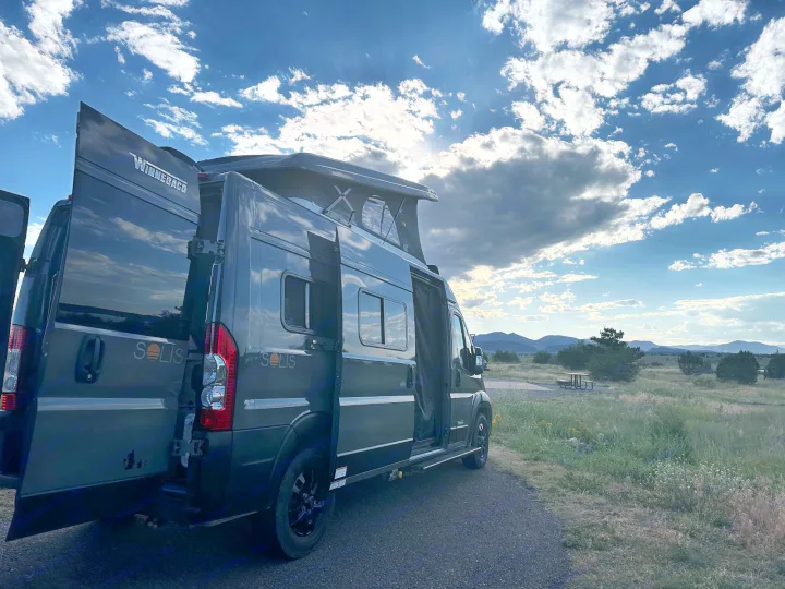 Dark gray Winnebago Solis rv rental van, pop-top up, doors open, in a scenic mountain landscape under a dramatic sky.. Winnebago Solis 2023