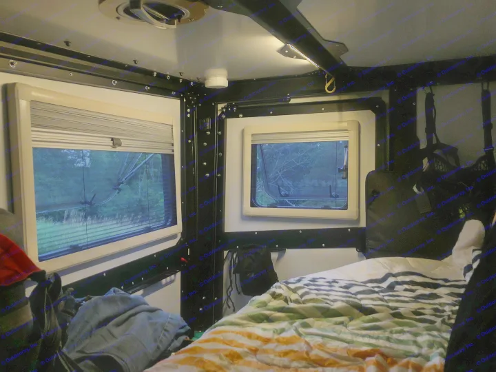 Cozy RV rental interior at dusk: two windows reveal a wooded view, complemented by a comfortable bed and soft ceiling lighting.. TAXA TAXA Trailer 2019