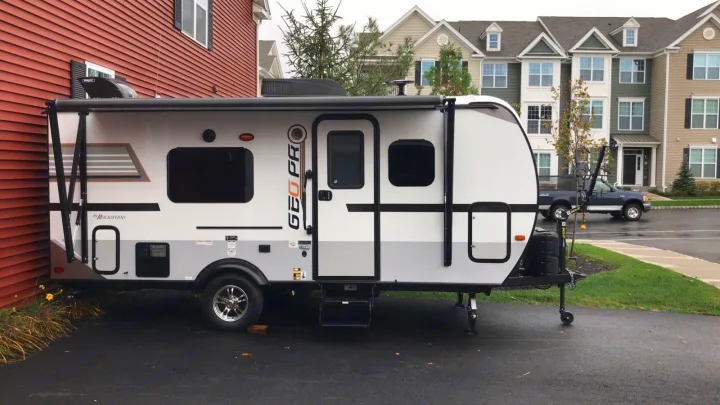 A sleek white and gray travel trailer (RV rental) parked on a driveway next to a red house with apartments in the background.. Forest River Rockwood 2018
