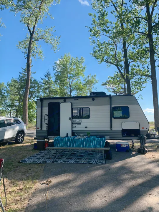 An RV rental parked at a sunny campsite with a picnic table setup and an SUV, surrounded by tall trees.. KZ Sportsmen classic 160QB 2021