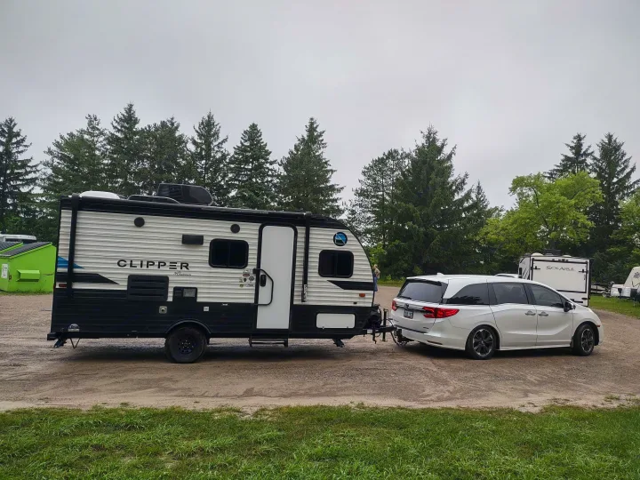 A white minivan tows a Clipper travel trailer RV at a campsite under an overcast sky. This setup is common for RV rental trips.. Coachmen Clipper 2020