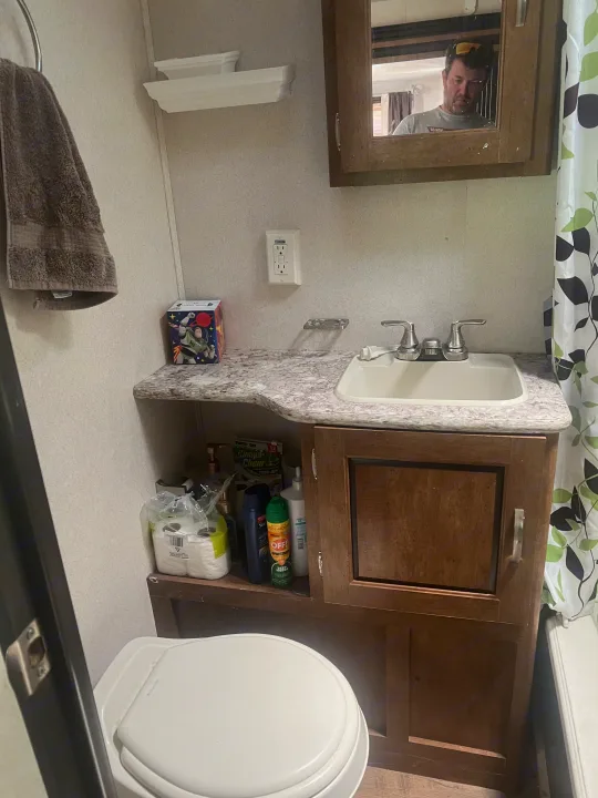 Inside a compact RV rental bathroom, showing the toilet, sink, mirror reflecting a man, and a leafy shower curtain.. Forest River Wildwood 2018