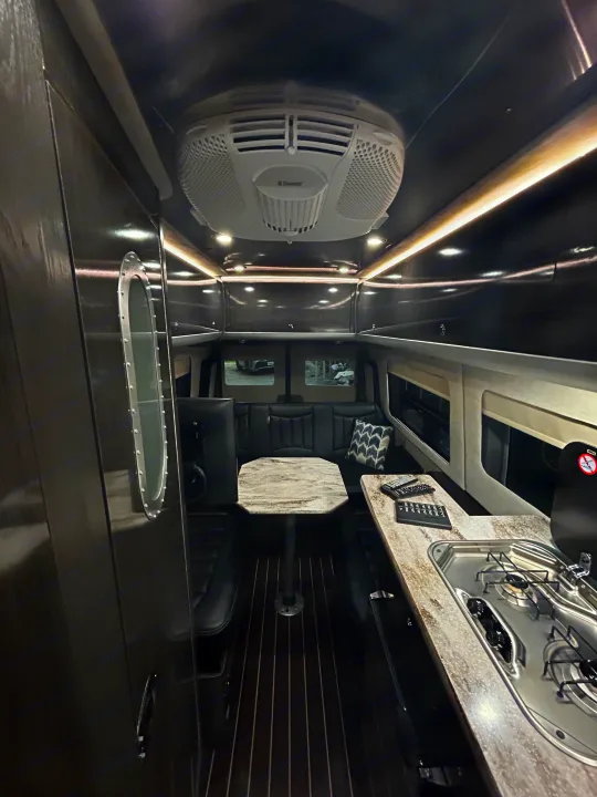 Explore this modern RV rental interior: cozy dining, kitchen with stovetop/sink, and overhead AC unit. Ready for travel!. Airstream Interstate 2016