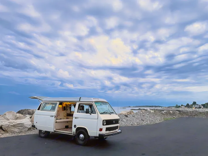 White VW camper van with open doors & cozy string lights, parked by a rocky coast. Perfect RV rental for your next adventure.. Volkswagen Westfalia 1983