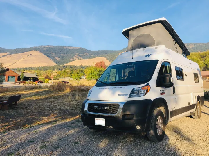 This white Winnebago Revel RV rental, with its pop-top extended, is parked on gravel, set against scenic mountains.. Winnebago Solis 2021