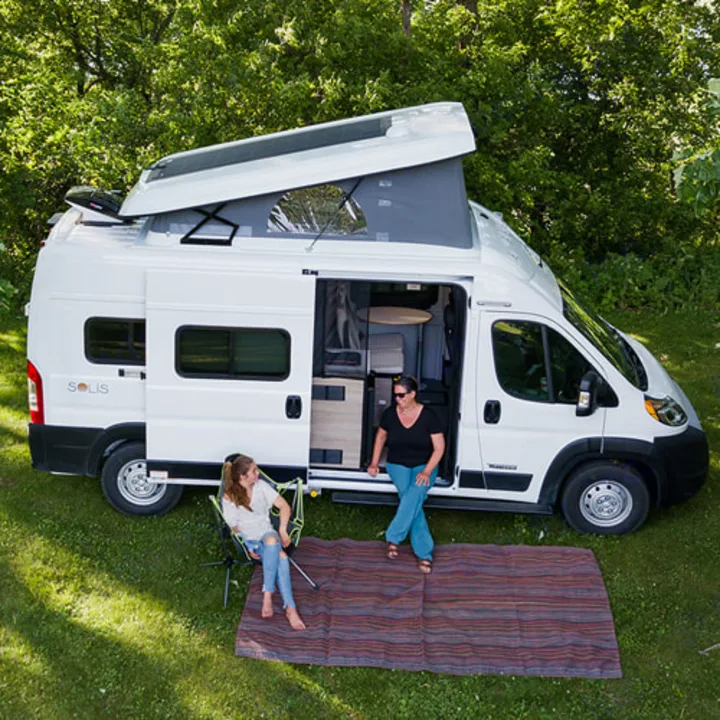 Enjoying an RV rental adventure! Two women relax by their white pop-top camper van in a grassy outdoor setting.. Winnebago Solis 2021