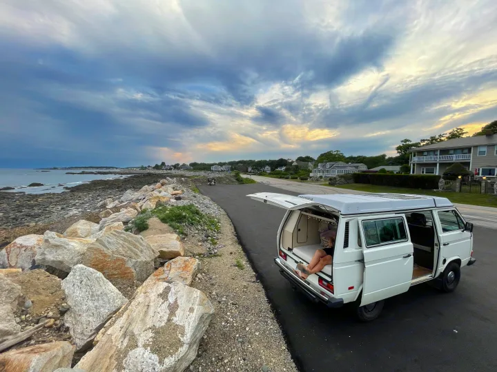 Person relaxing inside a classic white RV rental van overlooking a rocky coast & ocean.. Volkswagen Westfalia 1983