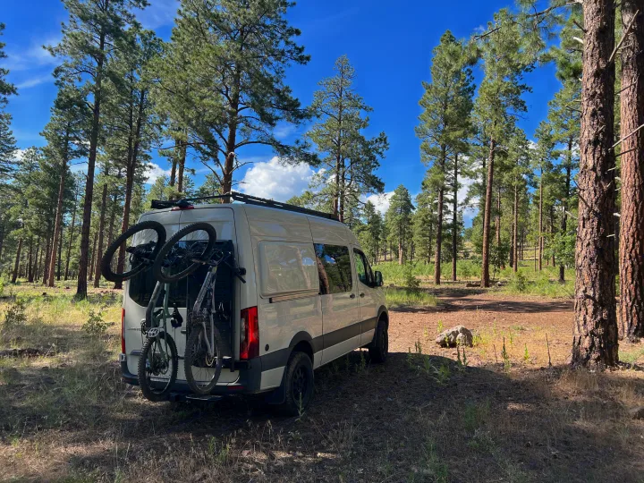White RV rental van, bikes on back, parked in a sunlit pine forest. Perfect adventure trip.. Mercedes-Benz Sprinter 2020