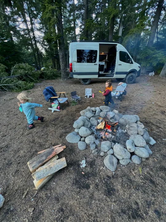 An RV rental is parked at a wooded campsite, where two young children play near a large, stone-ringed campfire.. Mercedes-Benz Sprinter 2020