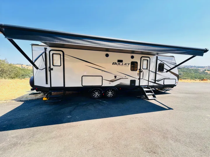 White travel trailer RV rental with an extended black awning parked on asphalt under a clear blue sky.. Keystone RV Bullet Ultra Lite 2023