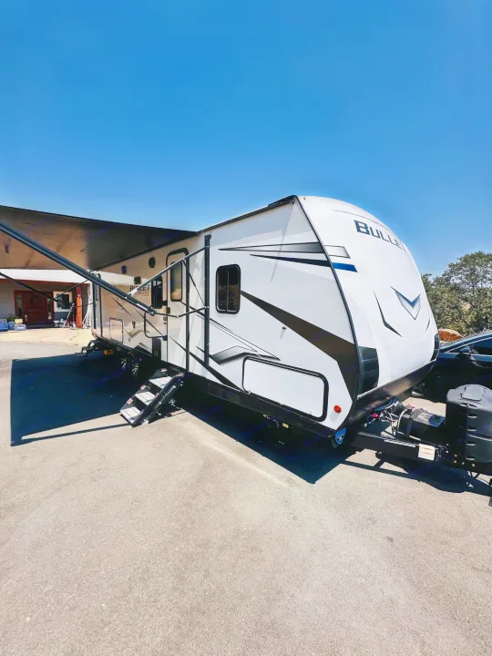 Modern white RV rental with extended awning and steps, parked under a clear blue sky, inviting outdoor adventures.. Keystone RV Bullet Ultra Lite 2023