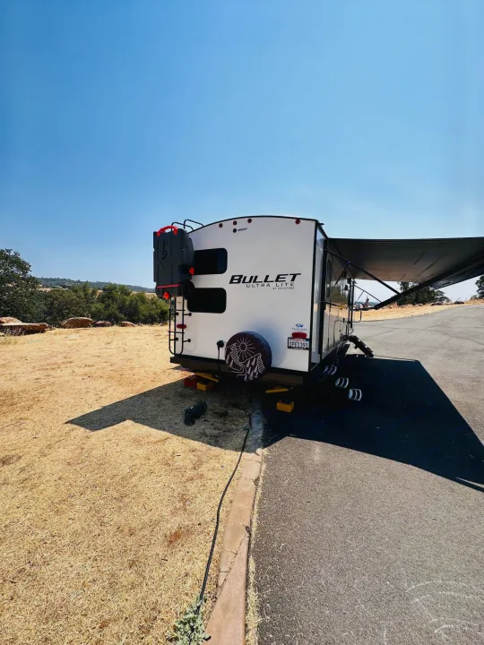 A white Bullet travel trailer RV rental with an extended awning is parked on asphalt next to dry grass, ready for camping on a clear, sunny day.. Keystone RV Bullet Ultra Lite 2023