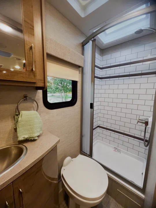 Clean RV rental bathroom with a skylit shower, subway tiles, toilet, vanity, and window showing trees outside.. Entegra Coach Other 2023