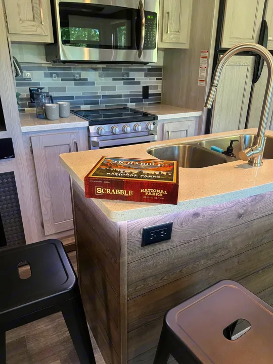 RV rental kitchen interior: Scrabble game on light countertop, stainless appliances, tile backsplash, and bar stools.. Keystone RV Montana High Country 2020